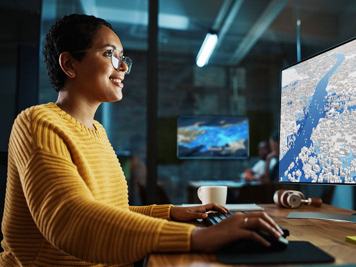 A woman in a yellow sweater uses a computer mouse and keyboard to navigate a 3D map displayed on a computer screen.