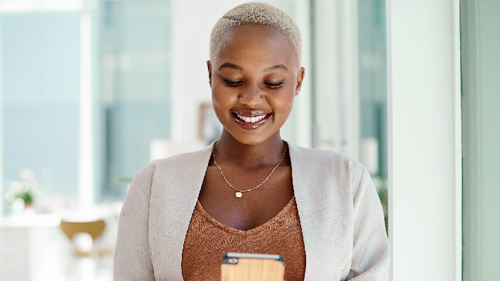 A smiling person wearing a pale jacket smiling down at a mobile phone in their hands with a sleek modern glass office
