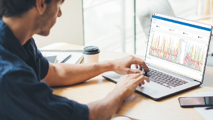 A person wearing a dark blue collared shirt seated at a desk using an ArcGIS Monitor interface displaying metrics on a small laptop