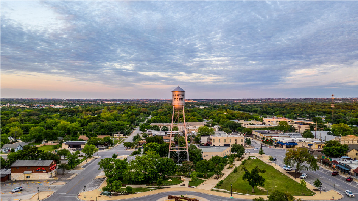 Aerial image of a water tower in the center of a neat town filled with green trees and small brown buildings under a cloud-streaked sunset sky 
