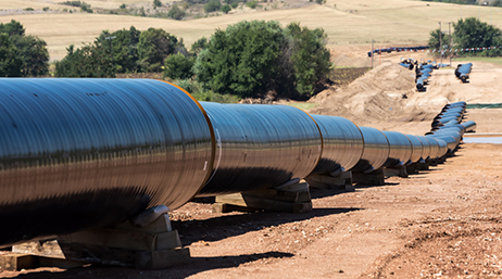 A long above-ground pipeline running across dry, brown land with green trees in the distance