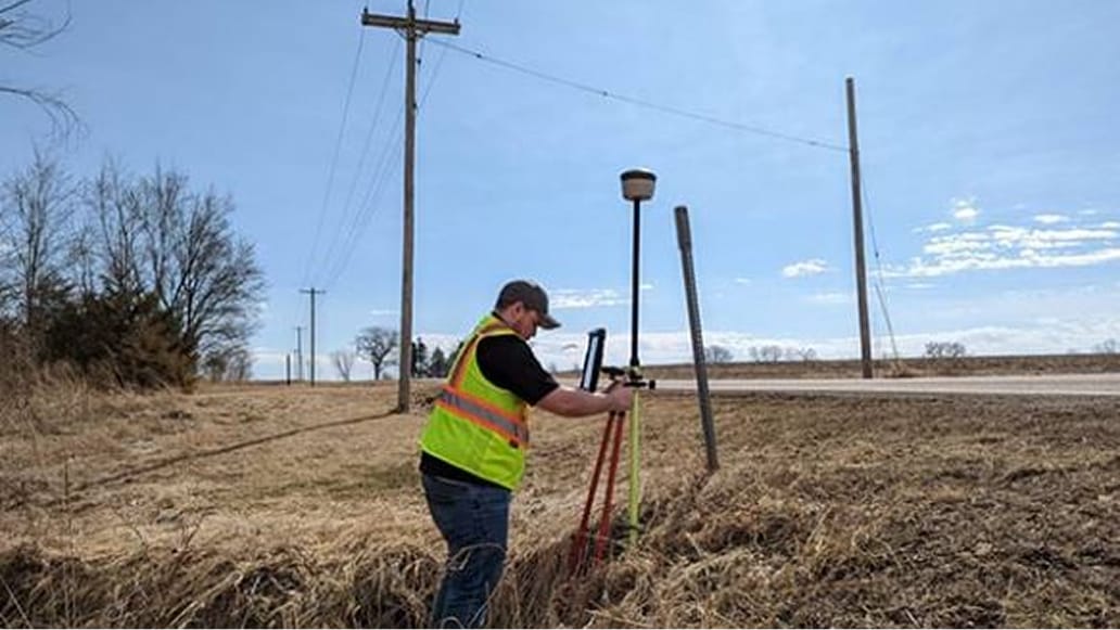 A person wearing a yellow safety vest setting up a tripod in a large field covered in thick brown brush