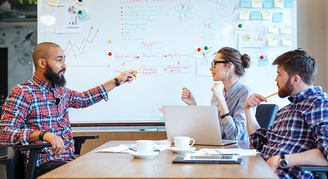 Three people sitting around a conference table discussing a large whiteboard covered with charts and graphs