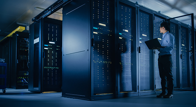 : A professionally dressed person carrying a laptop inspecting a large storage bank in a dimly lit room