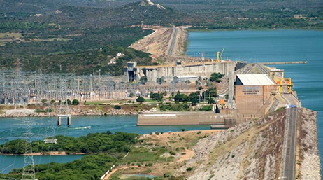 An aerial view of a dam with rippling blue water on one side and tree-dotted fields on the other