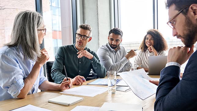 People gathered around a table in a conference room talking and viewing paper reports