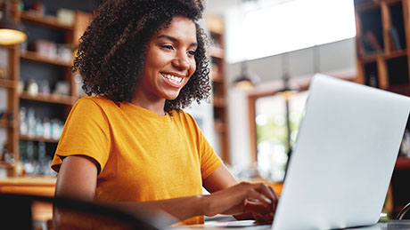 A photo of a smiling teenage student in a yellow shirt working on a laptop in a library setting