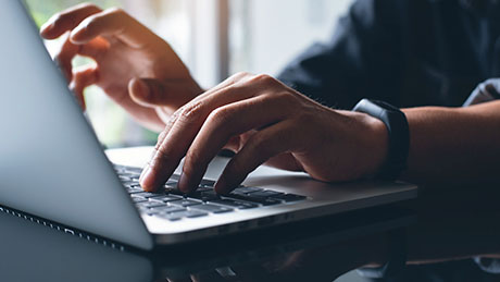 A closeup photo of a pair of hands wearing a watch typing on a laptop keyboard in a home office environment