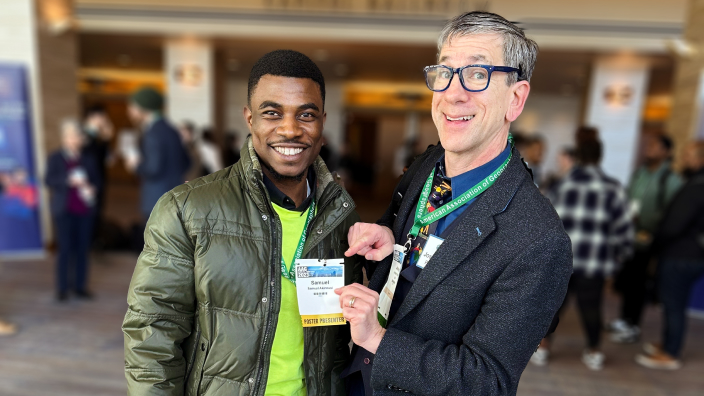 A student presenter wearing a conference badge poses with a GIS professional in a crowded expo hall