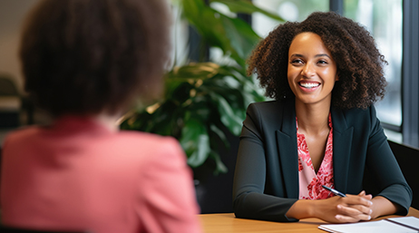 A smiling interviewer in a gray blazer and pink blouse faces an interviewee in a sunlit modern office