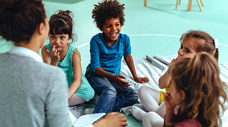 A photo of four laughing children and an adult sitting in a circle on the floor of a colorful classroom