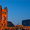 A photo of a city bridge lit up for nighttime reflecting the orange light of sunrise against a dark blue cityscape under a clear dark blue sky