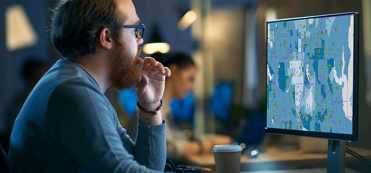 A person in a long sleeved blue tee shirt sits at a desk in a shared office space, using a monitor to access a digital map