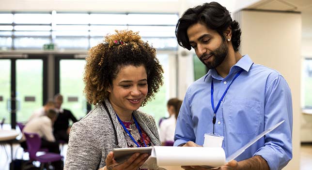 Two smiling teachers in blue lanyards standing in a school common area comparing data from a tablet and a notepad of paper