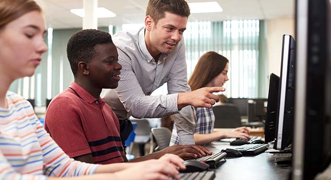 Three students sitting in a row at computer stations in a brightly lit modern classroom as a teacher leans between them to explain one of the screen displays