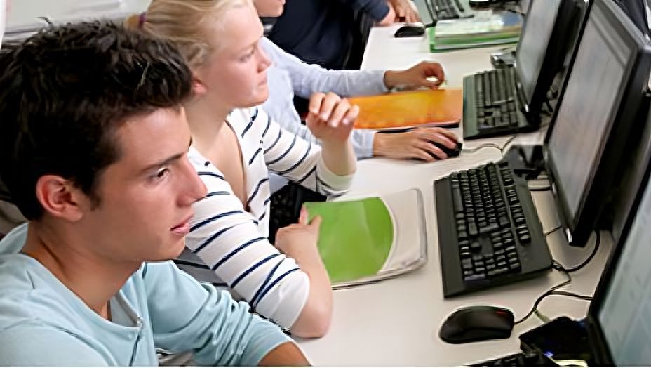 A row of students seated at school computer stations in a brightly lit modern classroom