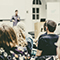 The backs of peoples’ heads as they sit in in a lecture hall and listen to a speaker standing in front of them in soft focus