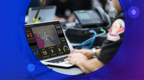 A first responder working in a room full of other people, and this first responder is tying on a laptop displaying a dashboard 