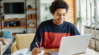 A smiling teenager sitting at a table with a laptop, a pen, and a notebook